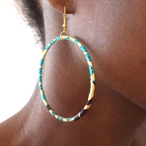 Peut inclure: Grandes boucles d'oreilles créoles dorées avec un tissu à motifs turquoise, noir et jaune enroulé autour des cerceaux. Les boucles d'oreilles sont larges et circulaires, avec une simple fermeture à crochet. Un accessoire audacieux et coloré.