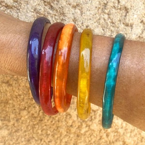 Puede incluir: Cinco pulseras de colores apiladas en la muñeca de una persona. Las pulseras son de color morado, rojo, naranja, amarillo y azul verdoso.
