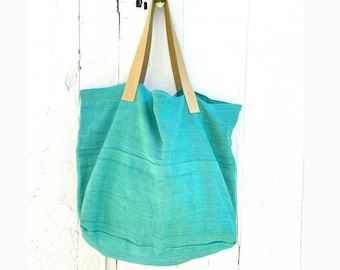 Puede incluir: Una bolsa de lona de color turquesa con asas de cuero marrón. La bolsa está colgada de una puerta de madera blanca.