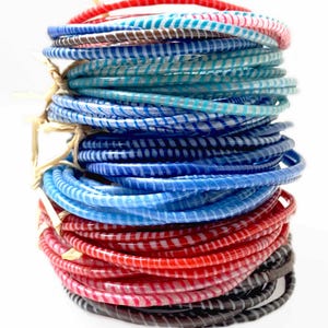 Peut inclure: Une pile verticale de bracelets fins et circulaires présentant une base translucide avec un motif de rayures blanches en spirale. Les bracelets sont disposés dans un dégradé de couleurs incluant le rouge, le bleu clair, le bleu foncé et le noir, liés par du raphia.