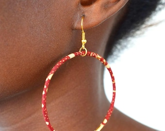 May include: A large circular hoop earring featuring a fabric-wrapped design in deep red with small, irregular cream-colored speckles. The hoop is suspended from a thin gold-toned metal hook that attaches to the top of the circular frame.