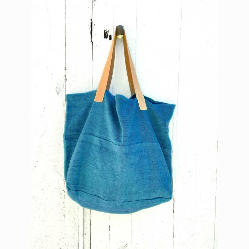 Puede incluir: Una bolsa de lona azul turquesa con asas de cuero marr&oacute;n colgando de una puerta de madera blanca.