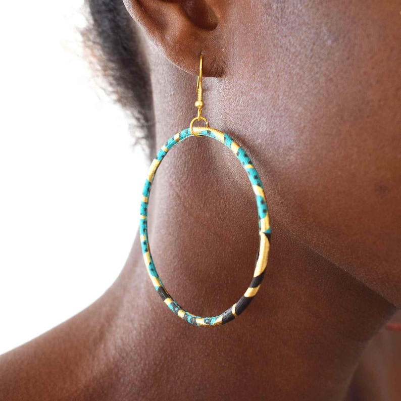 Peut inclure: Grandes boucles d'oreilles créoles dorées avec un tissu à motifs turquoise, noir et jaune enroulé autour des cerceaux. Les boucles d'oreilles sont larges et circulaires, avec une simple fermeture à crochet. Un accessoire audacieux et coloré.