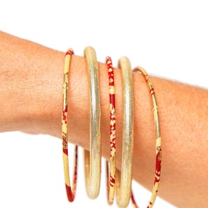 May include: A set of five gold bangles with red and gold patterned accents. The bangles are stacked on a person's wrist.