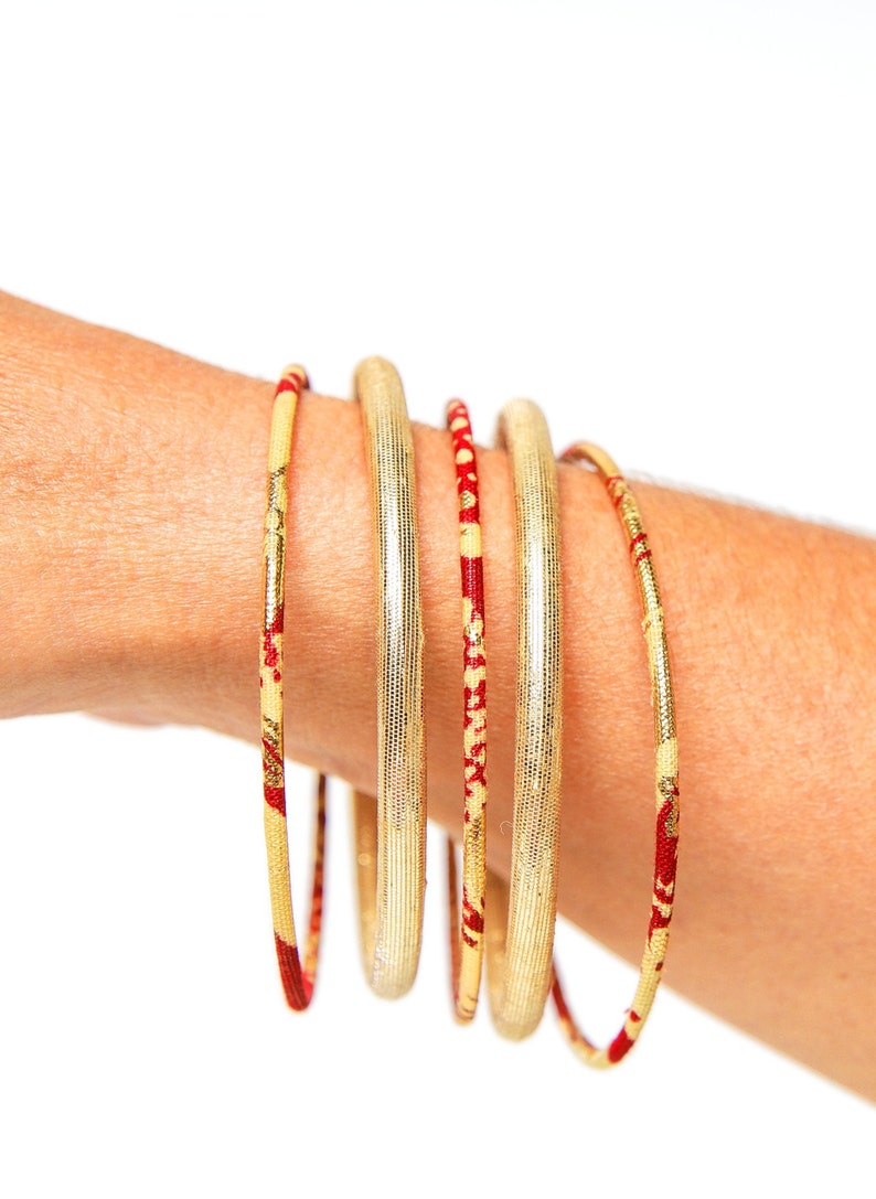 May include: A set of five gold bangles with red and gold patterned accents. The bangles are stacked on a person's wrist.