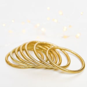 May include: A stack of gold-colored metal bangles. The bangles are smooth and shiny, and they are arranged in a loose pile.