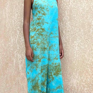 May include: A turquoise jumpsuit with a tie-dye pattern in shades of green and brown. The jumpsuit features thin straps and wide legs. The model is standing in front of a textured beige wall.