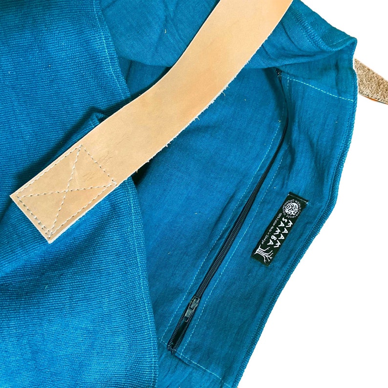 Puede incluir: Una bolsa de lona azul turquesa con una correa de cuero marr&oacute;n y un bolsillo con cremallera. La bolsa tiene una etiqueta negra con el texto "YAAAY" y un dise&ntilde;o de &aacute;rbol.