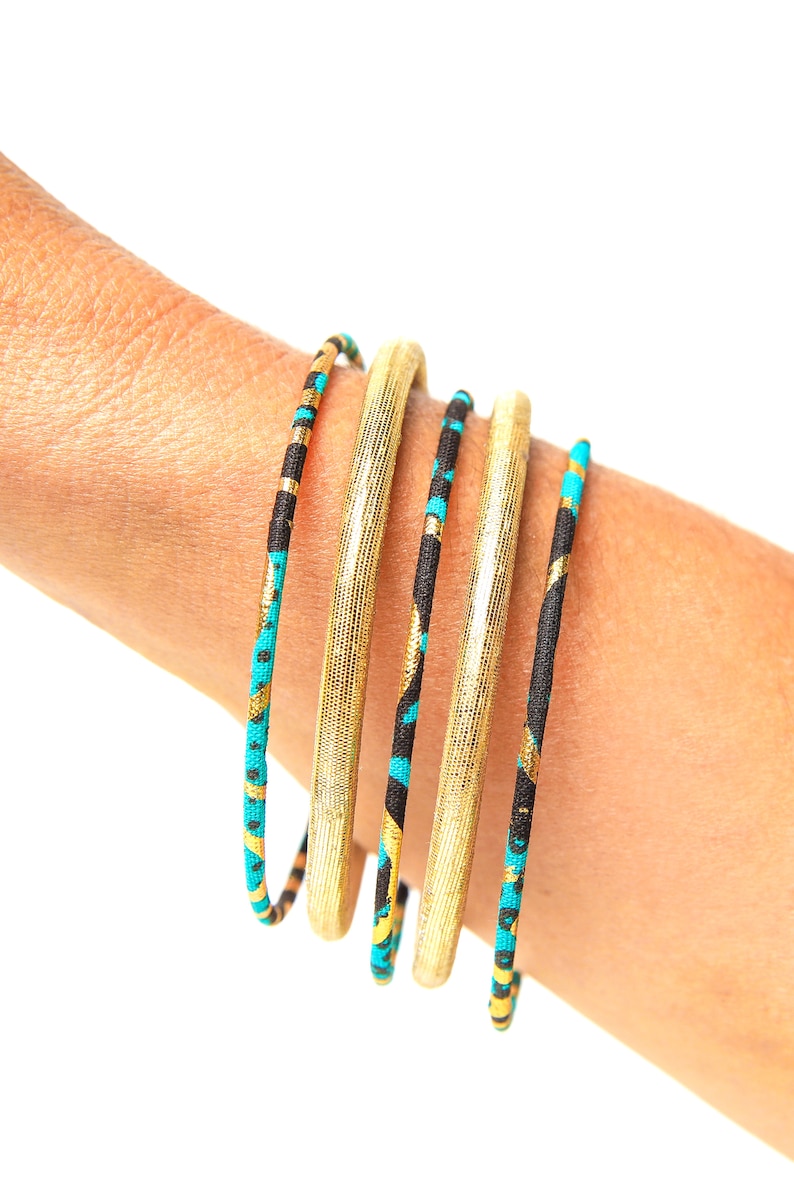 May include: Five gold and black bangles with turquoise accents. The bangles are stacked on a person's wrist.