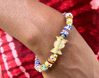 May include: A colorful beaded bracelet with a gold-colored centerpiece. The bracelet features a mix of cylindrical and round beads in various colors, including blue, yellow, red, and green. The gold centerpiece has a detailed, ornate design.