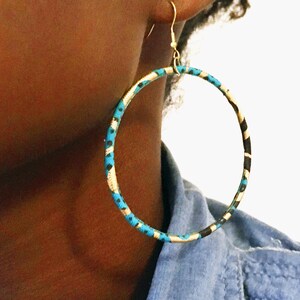 May include: Large hoop earrings with a colorful fabric wrap. The earrings feature a gold-colored metal hook and a circular hoop design. The fabric wrap has a pattern of blue, black, and gold. The earrings are worn on a person's ear.