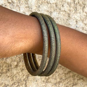 Könnte beinhalten: Drei goldfarbene Armreifen mit strukturierter Oberfläche sind auf einem Handgelenk gestapelt. Die Armreifen haben einen metallischen Glanz, der das Licht reflektiert. Der Hintergrund ist eine neutrale, strukturierte Oberfläche.