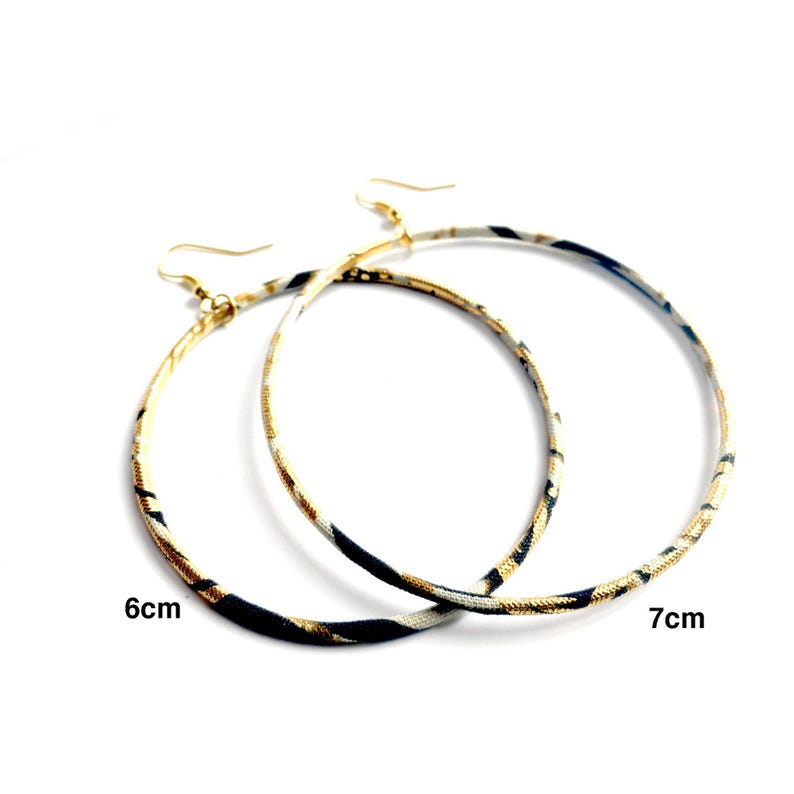 May include: Two gold-tone hoop earrings. The hoops are covered in a fabric with a black, white, and gold pattern. One earring is 6 cm in diameter, and the other is 7 cm in diameter. The earrings have gold-tone ear wires.