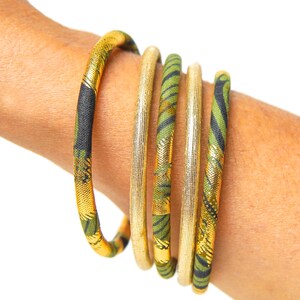 May include: Five gold and green fabric bangles stacked on a person's wrist. The bangles have a geometric pattern and are accented with gold thread.