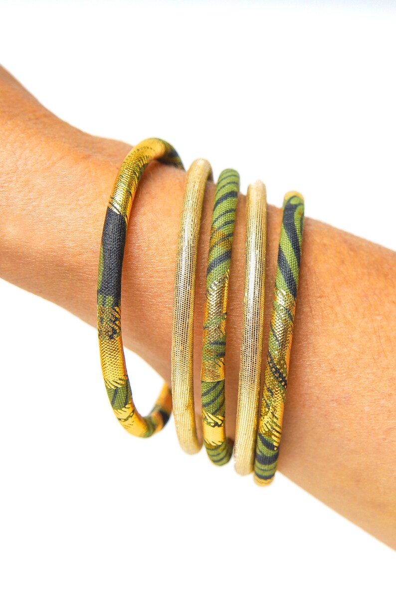 May include: Five gold and green fabric bangles stacked on a person's wrist. The bangles have a geometric pattern and are accented with gold thread.