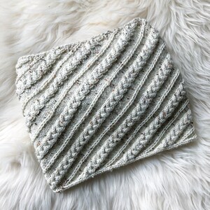 May include: A white knitted cowl with a diagonal cable pattern. The cowl is made with a speckled yarn that has a subtle fleck of brown.