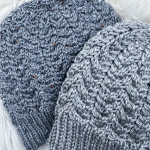 Könnte beinhalten: Zwei graue Strickmützen mit unterschiedlichen Strickmustern. Die Mützen sind aus einem weichen, strukturierten Garn gefertigt.