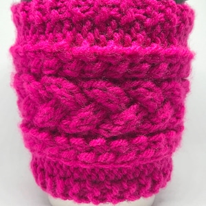 May include: A bright pink knitted coffee cup cozy with a cable knit pattern.