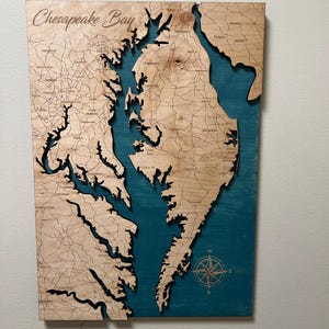 May include: Wooden map of the Chesapeake Bay with engraved details of the coastline, roads, and cities. The bay area is painted teal, and the text "Chesapeake Bay" is at the top. A compass rose is in the lower right corner.