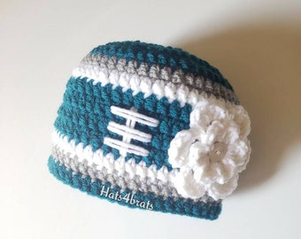Puede incluir: Sombrero de punto de color turquesa, gris y blanco con una flor blanca de ganchillo. El sombrero tiene una banda blanca con un diseño cosido en la parte delantera. El sombrero está etiquetado como "Hats4brats".
