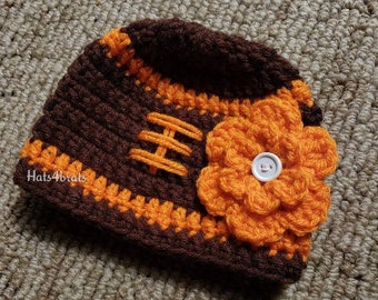 May include: A brown and orange crocheted hat with a football design and a large orange crocheted flower with a white button in the center. The hat is on a brown carpet.