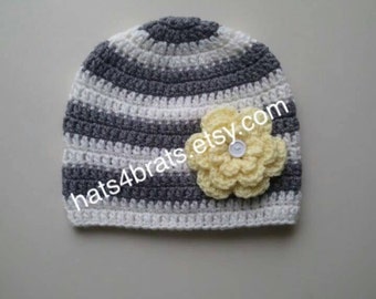 Op de afbeelding: Grijze en witte gestreepte gebreide beanie met een gele gehaakte bloem en een knoop in het midden. De beanie heeft de tekst "hats4brats.etsy.com" aan de zijkant.
