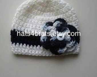 Op de afbeelding: Witte gehaakte baby muts met een zwarte en blauwe bloem en een witte knoop in het midden. De muts heeft een zwarte streep rond de onderkant. De website hats4brats.etsy.com is zichtbaar op de muts.