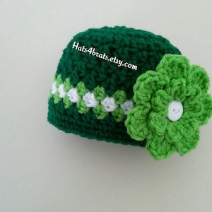 Puede incluir: Sombrero de bebé de ganchillo verde con una franja blanca y verde y una flor de ganchillo verde con un botón blanco en el centro. El sombrero está a la venta en Etsy en hats4brats.etsy.com.