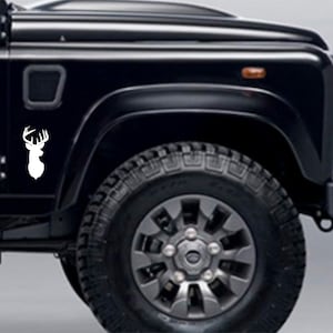 May include: A black Land Rover Defender with a white deer head silhouette decal on the front fender. The vehicle has a black tire with a silver rim.