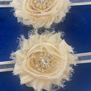 May include: Two ivory fabric flower hair accessories with a rhinestone center. The flowers are attached to a white and blue striped headband. The background is a solid blue.