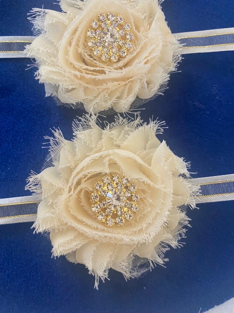 May include: Two ivory fabric flower hair accessories with a rhinestone center. The flowers are attached to a white and blue striped headband. The background is a solid blue.