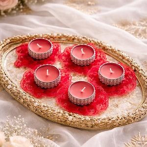 May include: An ornate gold-coloured tray displays five red candles with silver rims, each on a red lace doily. The tray is set against white fabric and floral decorations, creating an elegant arrangement.