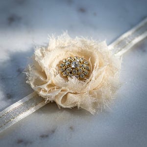 May include: Ivory fabric flower with a gold and rhinestone center. The flower is attached to a thin, shimmering ribbon. The petals have a frayed edge, giving it a vintage or handmade appearance. The design is delicate and elegant.