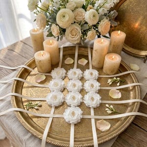 May include: A decorative arrangement featuring a gold-toned tray with white floral accents and candles. The tray holds white floral embellishments with gold centers, complemented by ivory ribbons, lit candles, and a bouquet of white flowers.