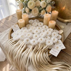 May include: A decorative arrangement featuring white floral accents, lit candles, and a bouquet of white flowers. The display is set on a gold-toned tray with long, cream-colored ribbons. Small cards are also present.