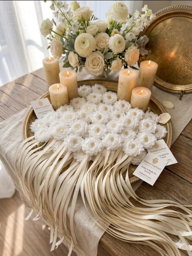May include: A decorative arrangement featuring white floral accents, lit candles, and a bouquet of white flowers. The display is set on a gold-toned tray with long, cream-colored ribbons. Small cards are also present.