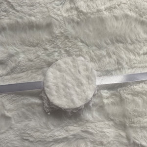 May include: A white, circular, fluffy object with a delicate lace trim, attached to a thin, white satin ribbon. The object is set against a soft, white, textured background.