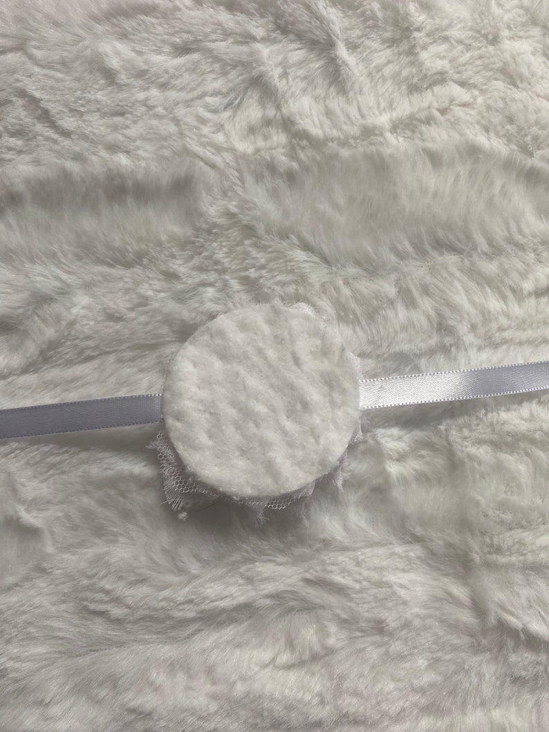 May include: A white, circular, fluffy object with a delicate lace trim, attached to a thin, white satin ribbon. The object is set against a soft, white, textured background.