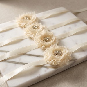 May include: Ivory-colored floral embellishments with rhinestone centers are attached to ivory satin ribbons. The flowers are made of layered fabric with a frayed edge. The ribbons are arranged on a white marble surface.