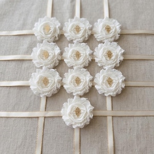 May include: A display of nine white fabric flowers with gold-colored Hamsa hand charms in the center. The flowers are arranged on a beige fabric background with cream-colored ribbons forming a grid pattern.