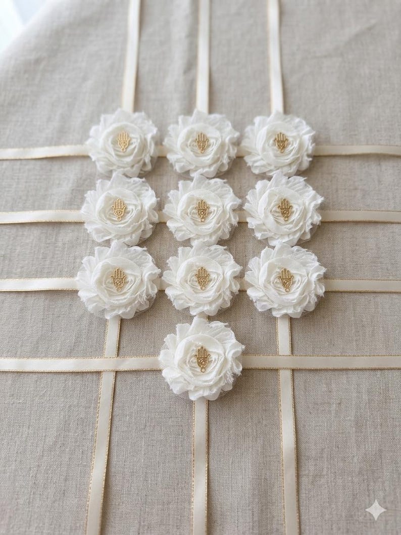May include: A display of nine white fabric flowers with gold-colored Hamsa hand charms in the center. The flowers are arranged on a beige fabric background with cream-colored ribbons forming a grid pattern.