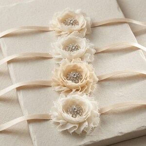 May include: Four ivory fabric flower embellishments with rhinestone centers and attached cream-colored satin ribbons. The flowers are layered with ruffled edges, creating a delicate, textured appearance. Ideal for wedding or craft projects.