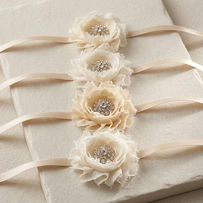 May include: Four ivory fabric flower embellishments with rhinestone centers and attached cream-colored satin ribbons. The flowers are layered with ruffled edges, creating a delicate, textured appearance. Ideal for wedding or craft projects.