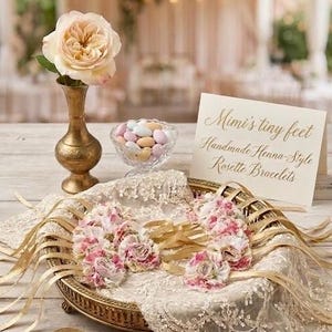 May include: A still life arrangement featuring a peach-colored rose in a gold vase, a glass bowl of candies, and a decorative tray with rosette bracelets. The tray is adorned with lace and gold ribbons. A sign reads "Mimi's tiny feet".