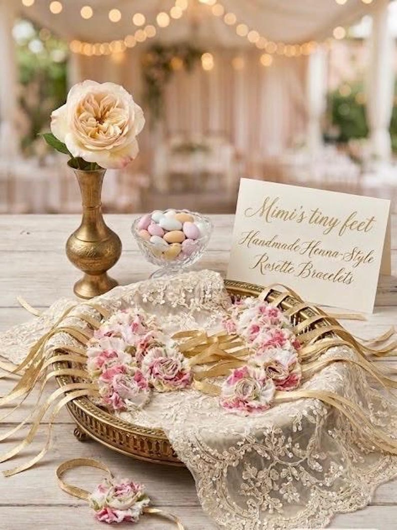 May include: A still life arrangement featuring a peach-colored rose in a gold vase, a glass bowl of candies, and a decorative tray with rosette bracelets. The tray is adorned with lace and gold ribbons. A sign reads "Mimi's tiny feet".