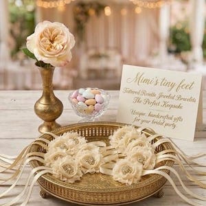 May include: A gold-toned decorative tray holds several ivory rosette bracelets with ribbon accents. A small glass bowl of pastel candies and a vase with a peach rose are also present. The text on the card reads "Mimi's tiny feet".