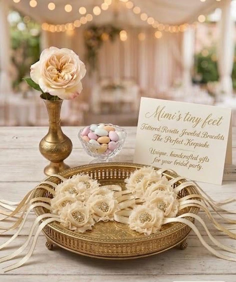 May include: A gold-toned decorative tray holds several ivory rosette bracelets with ribbon accents. A small glass bowl of pastel candies and a vase with a peach rose are also present. The text on the card reads "Mimi's tiny feet".