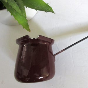 May include: A small, dark brown enamel coffee pot with a long, thin handle and a silver ring at the end. The pot has a flared rim and a rounded body. The handle is attached to the side of the pot. The background is white.
