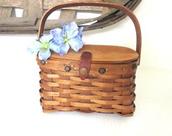 May include: A small, rectangular woven basket with a wooden lid and handle. The basket is made of light brown wood and has a leather strap closure. Two light blue flowers with green centres rest on top of the lid. The basket is a decorative item.