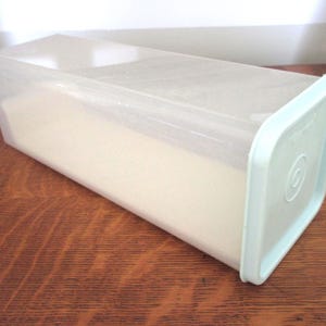 May include: A rectangular, clear plastic container with a pale blue lid. The container is filled with a white substance. The container is on a wooden surface.